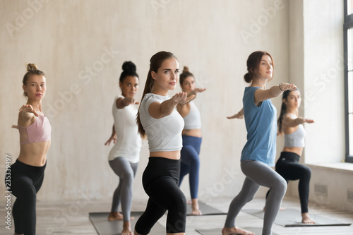 Group of young sporty people practicing yoga lesson, doing Warrior Two pose, Virabhadrasana 2 exercise, working out, indoor full length, mixed race students training at sport club or yoga studio photo