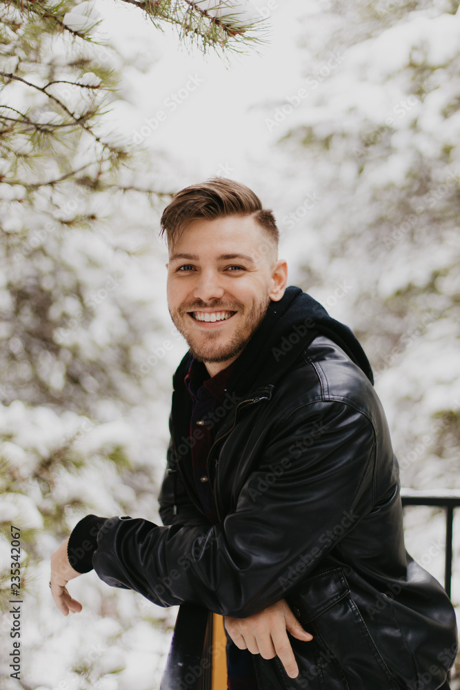 Portrait of Attractive Trendy Modern Young Male Model Smiling and Enjoying the Outdoors and the White Winter Snow in the Tree Canopy While Posing in Fashionable Leather Bomber Jacket Portrait of Attractive Trendy Modern Young Male Model Smiling and Enjoying the Outdoors and the White Winter Snow in the Tree Canopy While Posing in Fashionable Leather Bomber Jacket