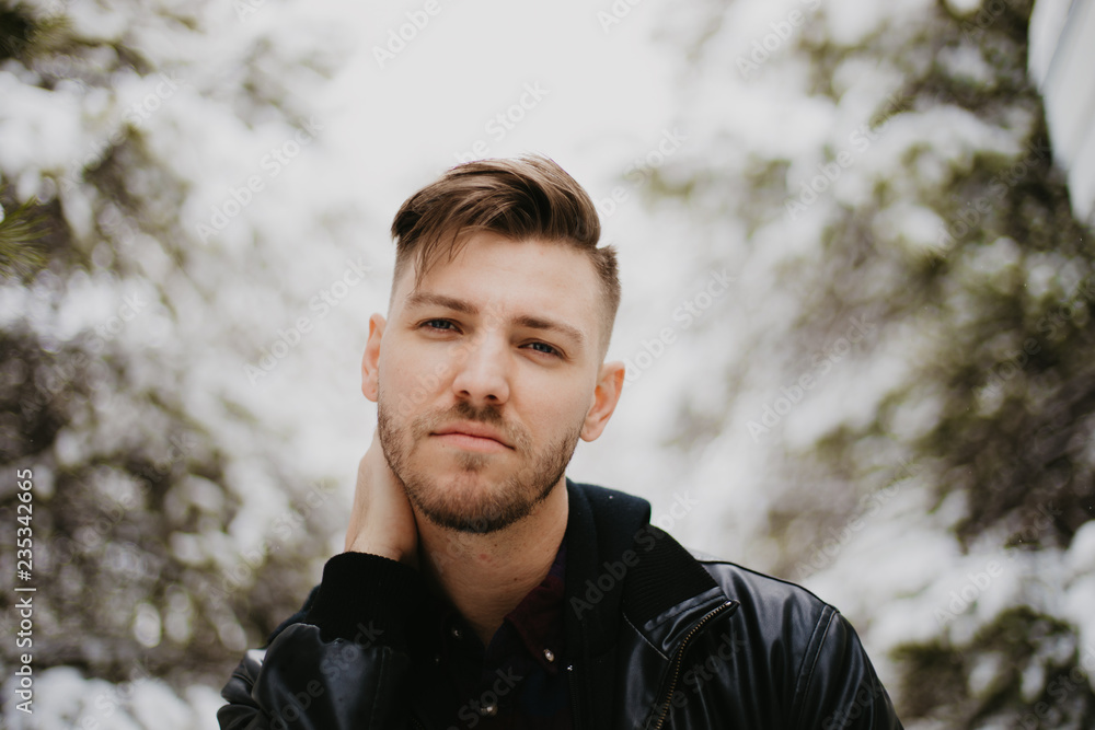 Portrait of Attractive Trendy Modern Young Male Model Smiling and Enjoying the Outdoors and the White Winter Snow in the Tree Canopy While Posing in Fashionable Leather Bomber Jacket Portrait of Attractive Trendy Modern Young Male Model Smiling and Enjoying the Outdoors and the White Winter Snow in the Tree Canopy While Posing in Fashionable Leather Bomber Jacket