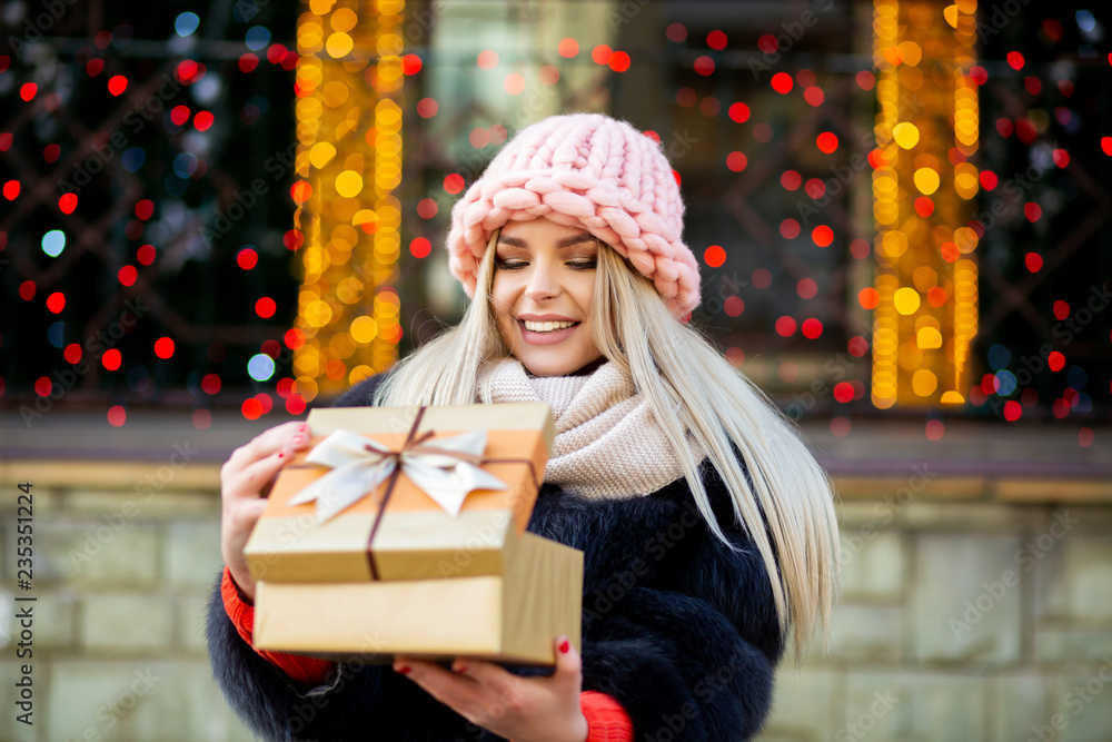 Cheerful blonde girl wearing knitted cap and warm coat, opening gift box at the background of city lights. Space for text