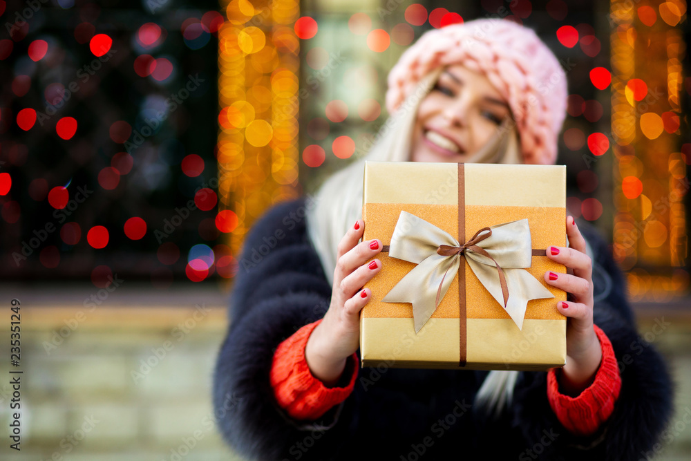 Charming blonde girl wearing fur coat and cap pulls a gift box to you. Space for text Charming blonde girl wearing fur coat and cap pulls a gift box to you. Space for text