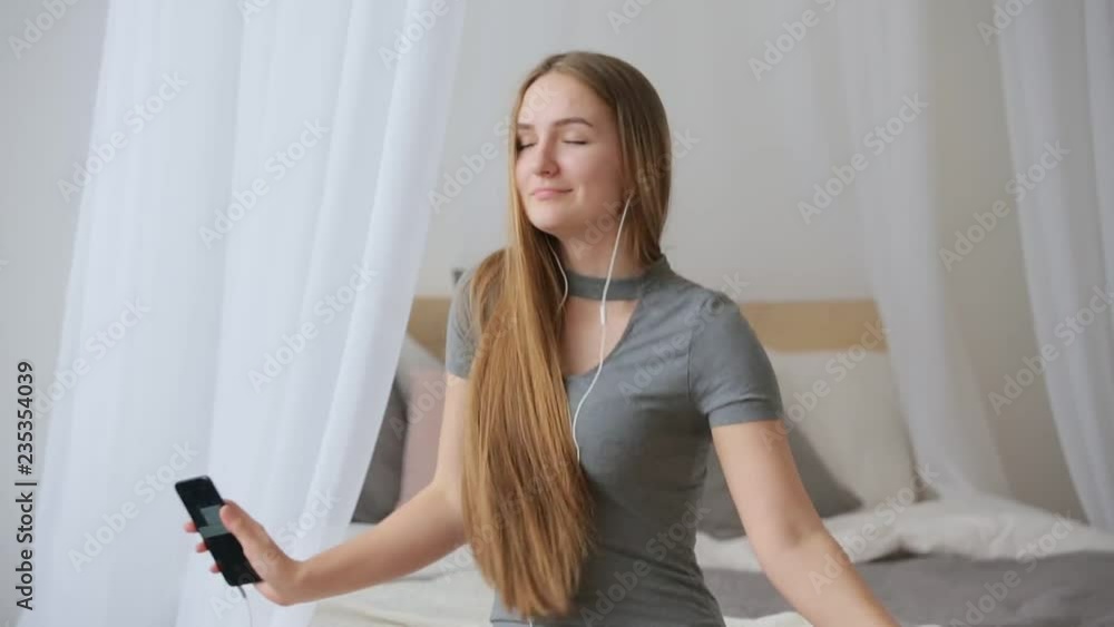Joyful young beautiful woman, girl in the bedroom sitting on the bed near the window and looks out the window and listening music on her smartphone. Joyful young beautiful woman, girl in the bedroom sitting on the bed near the window and looks out the window and listening music on her smartphone.