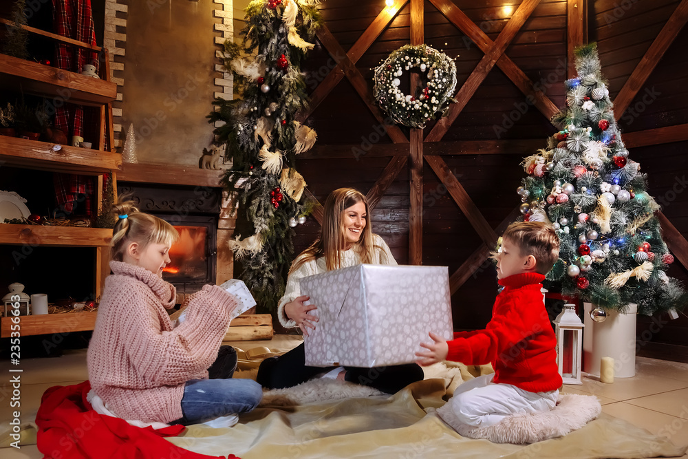 Merry Christmas and Happy New Year. Cheerful mom and her cute daughter and son exchanging gifts. Parent and children having fun near Christmas tree indoors. Loving family with presents in room.