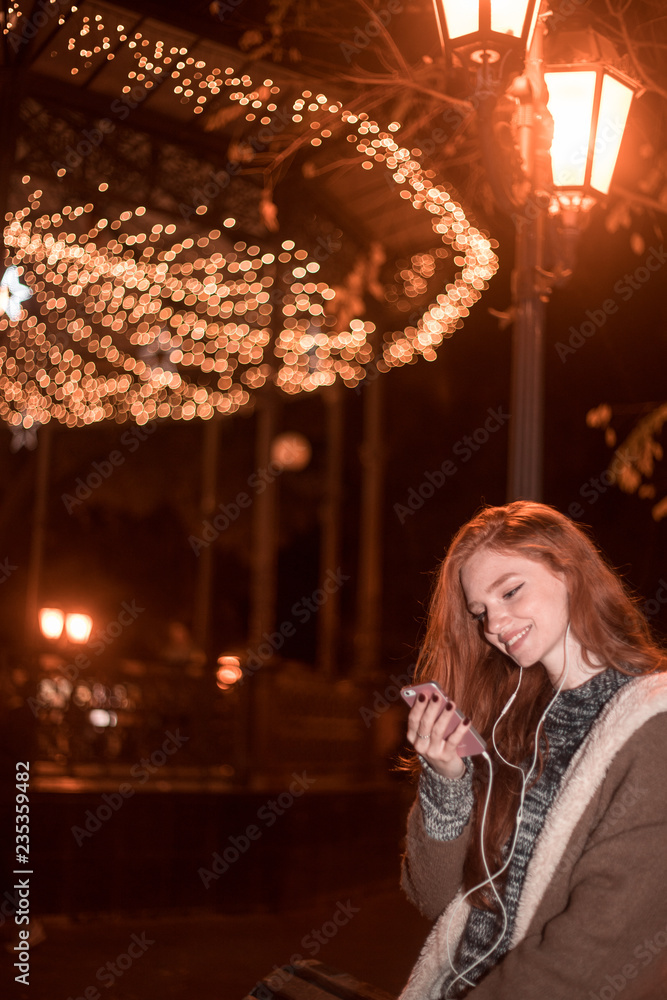 The portrait of an young smiling redhear lady on the evening listening musik watching her phone The portrait of an young smiling redhear lady on the evening listening musik watching her phone