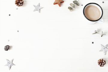 Christmas composition. Eucalyptus branches, wooden stars, pine cones, cinnamon sticks and glittering confetti on white wooden table background. Coffee in enamel mug. Winter breakfast scene. Flat lay