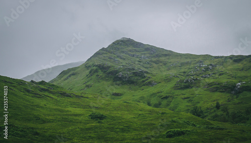 A green hill in the Scottish Highlands in the UK, surrounded by clouds and mist during a cold and rainy day.