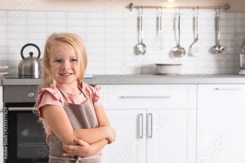 Cute little girl wearing apron near oven in kitchen. Space for text