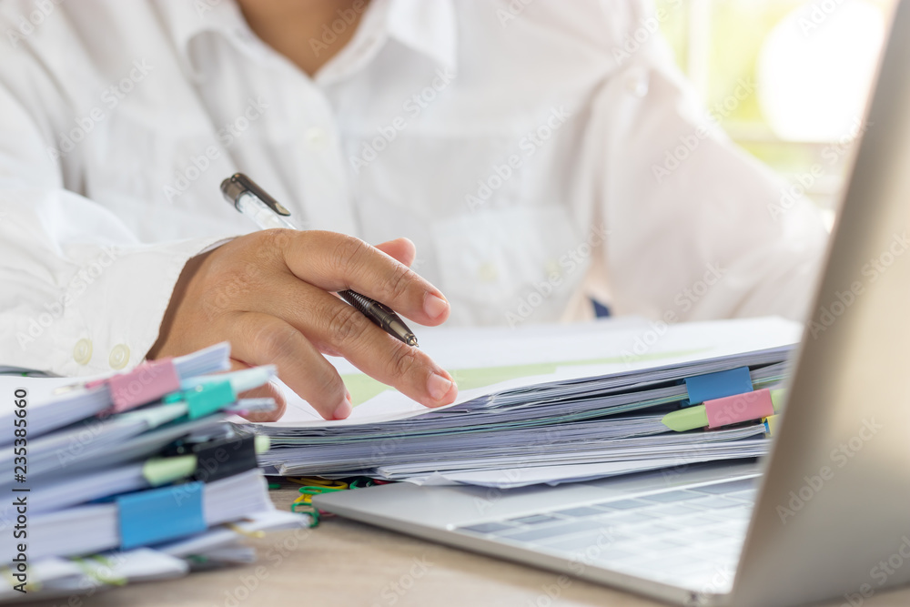 Teacher holding pen by hand for checking student homework assignments on desk in school office. Unfinished paperwork stacked in archive with color binder paper clips. Education and business concept. Teacher holding pen by hand for checking student homework assignments on desk in school office. Unfinished paperwork stacked in archive with color binder paper clips. Education and business concept.