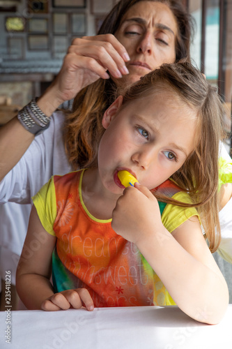 four years old blonde little cute girl eating ice lolly sitting on mother legs in restaurant