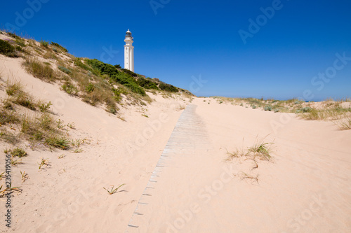 wooden footway on beach sand in nature next to the lighthouse of Cape Trafalgar, in Canos Meca village (Barbate, Cadiz, Andalusia, Spain), blue sky. Horizontal shot