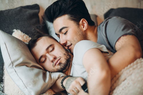 Tender handsome same sex male lovers, active young guy awakes his passive lover with a sweet kisss while sleeping in soft bed and hugging in the morning photo