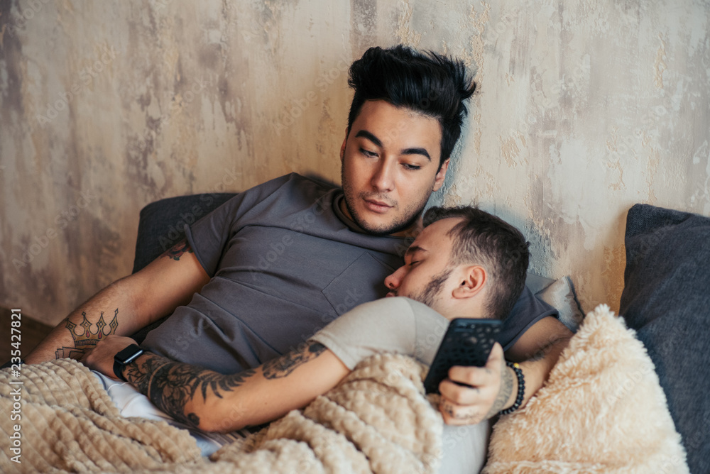 Foto Stock Alternative relationships, couple of young gay guys lying  together in bed and using smartphone to share music with each other. |  Adobe Stock