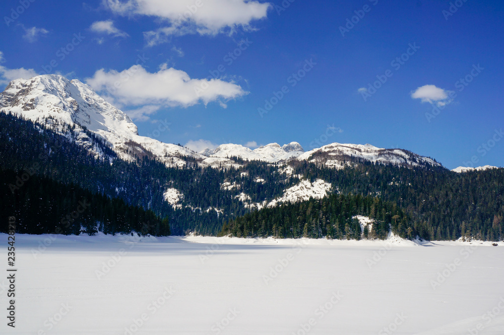 Winter landscape with a lake covered in untouched powder snow and tall mountain peaks in the background. Bobotov Kuk peak as seen from Black Lake (Crno jezero) in Zabljak, Montenegro.