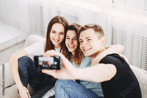 Carefree cheerful millennial friends taking group selfie on smartphone, happy excited young students making photo on cellphone hanging in living room together.
