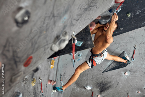 fitness, extreme hobbie, bouldering, people and healthy lifestyle concept - young positive male alpinist practicing climbing on indoor rock artificial boulder, equipped with belay ropes