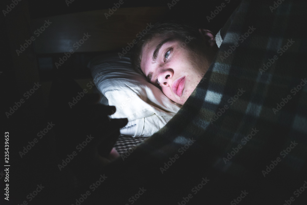Depressed teenager browsing the internet on his mobile phone as he is lying on his bed in the dark. Depressed teenager browsing the internet on his mobile phone as he is lying on his bed in the dark.
