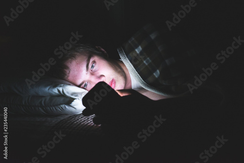 Depressed teenager browsing the internet on his mobile phone as he is lying on his bed in the dark.