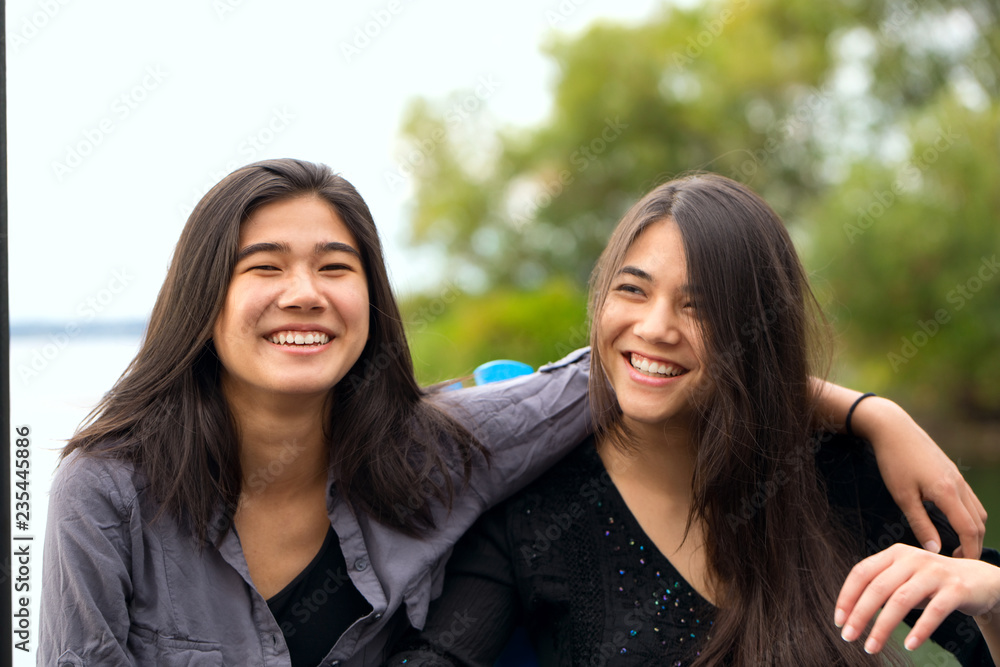Two smiling biracial teen girls smiling outdoors by lake, hugging Two smiling biracial teen girls smiling outdoors by lake, hugging