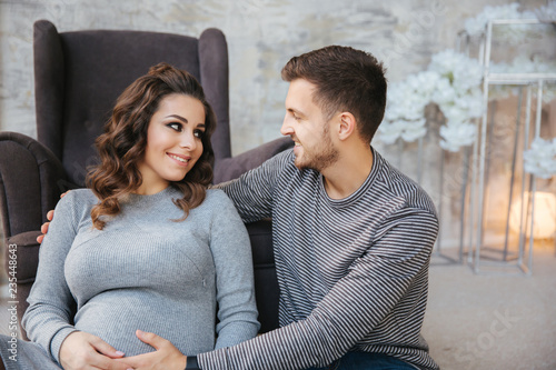 Young family sist near the chair and look at the belly. Pregman woman with her beloved man. Happy couple in Christmas mood photo