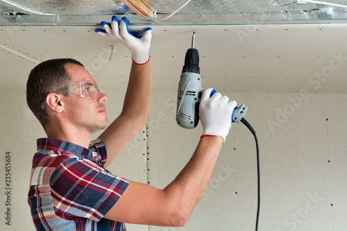 Young man in goggles fixing drywall suspended ceiling to metal frame using electrical screwdriver on ceiling insulated with shiny aluminum foil. Renovation, construction, do it yourself concept. photo