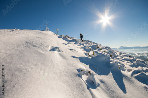 Tourist hiker climber in winter clothing with backpack climbing dangerous rocky steep mountain slope covered with deep snow, white sun rays shining in bright blue sky copy space background. © bilanol