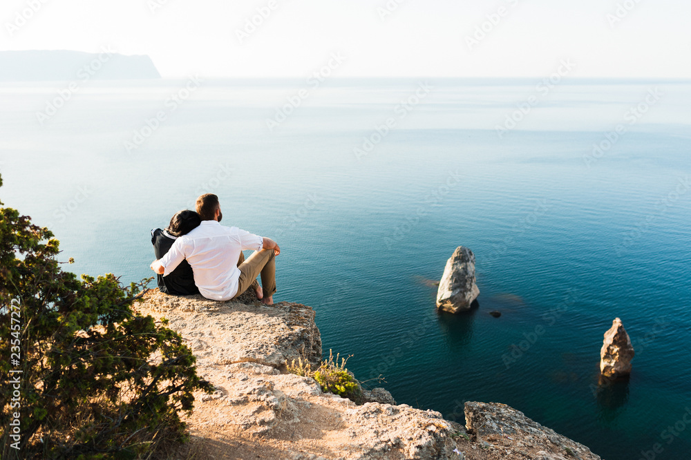Loving couple sitting on the edge of the cliff by the sea. Honeymoon. Honeymoon trip. Boy and girl at the sea. Man and woman traveling. Couple hugs. Couple kissing. Newly married couple. Lovers