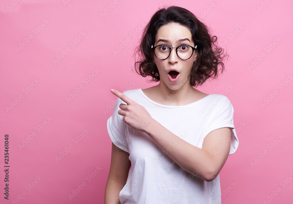 Good looking young Caucasian woman in round transparent eyewear, keeps hand raised, dressed in casual outfit, pretends holding something wonderful, isolated over pink background. Look there Good looking young Caucasian woman in round transparent eyewear, keeps hand raised, dressed in casual outfit, pretends holding something wonderful, isolated over pink background. Look there