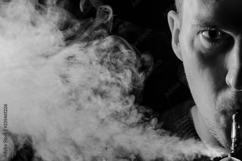 Portrait of a half part of guy with a shadow on a serious face  with a colored backlight of monochrome smoking a vape and exhaling white smoke in different directions on a black isolated background