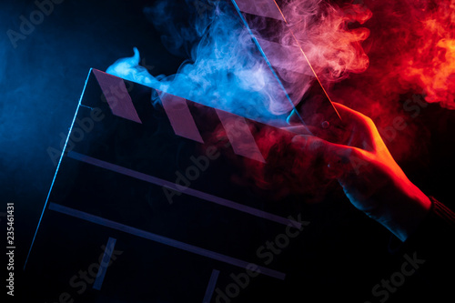 Close-up on an open clapperboard in hand before starting shooting a film with multi-colored smoke around with red and blue backlighting on a black isolated background