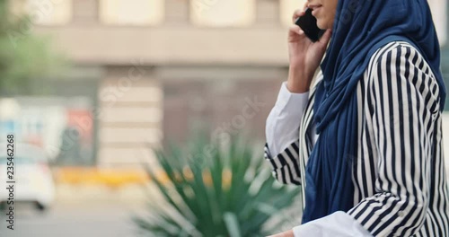 Attractive muslim girl wearing hijab walking down the street, talking with someone on phone and positively smiling - modern muslim, modern lifestyle concept closeup 4k