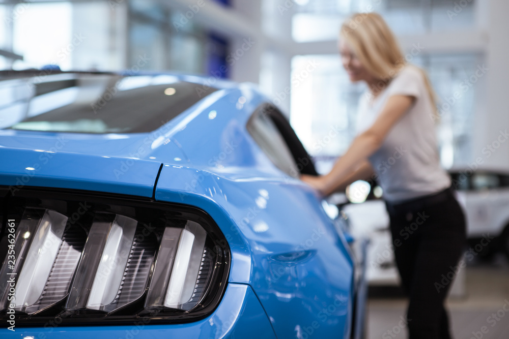 Selective focus on car lights on the foreground, woman looking inside the vehicle on the background, copy space. Female customer examining a car on sale at the dealership. Consumerism, driving concept Selective focus on car lights on the foreground, woman looking inside the vehicle on the background, copy space. Female customer examining a car on sale at the dealership. Consumerism, driving concept