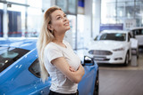 Charming young woman smiling happily, leaning on a new car at the dealership, copy space. Beautiful female customer looking away dreamily, choosing automobile on sale. Woman buying modern muscle car