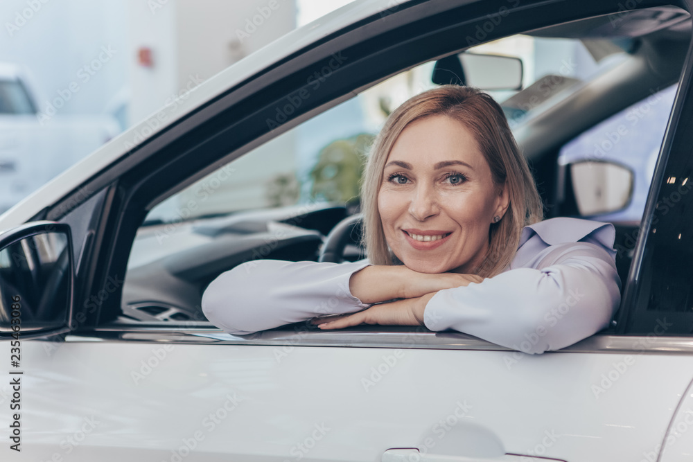 Mature cheerful woman sitting comfortably in a new automobile at the dealership salon, smiling happily to the camera. Mid-aged woman buying new car. Travelling, tourism, lifestyle concept