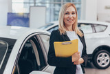 Professional car dealer posing proudly at auto showroom, smiling to the camera, holding clipboard. Happy saleswoman working at automobile dealership, copy space. Professionalism, management concept