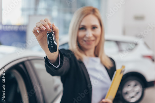 Car saleswoman smiling cheerfully, holding out car key to the camera, standing in front of new cars for sale. Selective focus on car keys in the hands of a female car dealer. Offer, discount concept