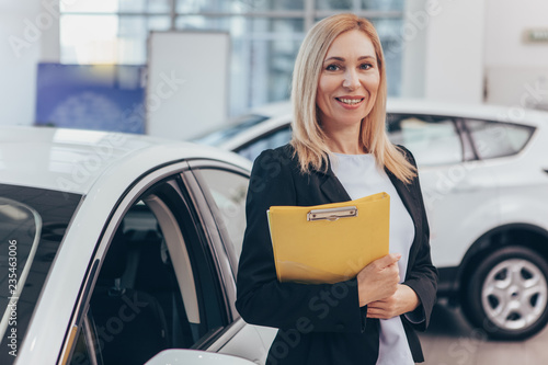 Foto Professional car dealer posing proudly at auto showroom, smiling to the camera, holding clipboard