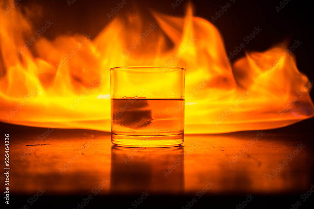 Whiskey in fire concept. Glass of whiskey and ice on wooden surface with color light and fog on background. Close up.