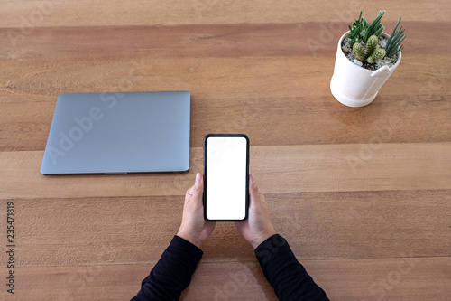 Top view mockup image of hands holding black mobile phone with blank desktop screen with laptop and cactus pot on wooden table background