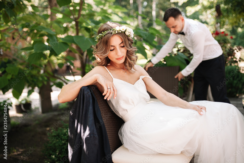 A beautiful bride in a white wedding dress and wreath sits on a chair next to the groom, resting and preparing for a happy family life A beautiful bride in a white wedding dress and wreath sits on a chair next to the groom, resting and preparing for a happy family life