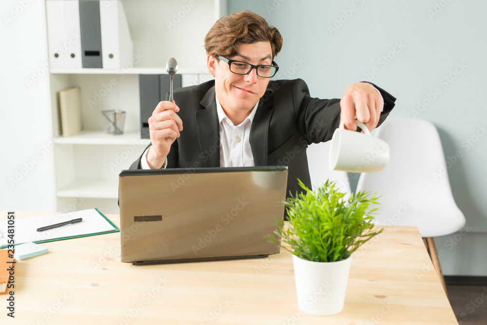 Office, humor, joke and business people concept - handsome man working in office, watering potted plant, sitting with spoon in his hands and smiling Office, humor, joke and business people concept - handsome man working in office, watering potted plant, sitting with spoon in his hands and smiling