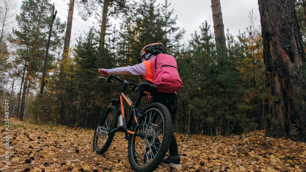 Naklejka premium One caucasian children walk with bike in autumn park. Little girl walking black orange cycle in forest. Kid goes do bicycle sports. Biker motion ride with backpack and helmet. Mountain bike hardtail.