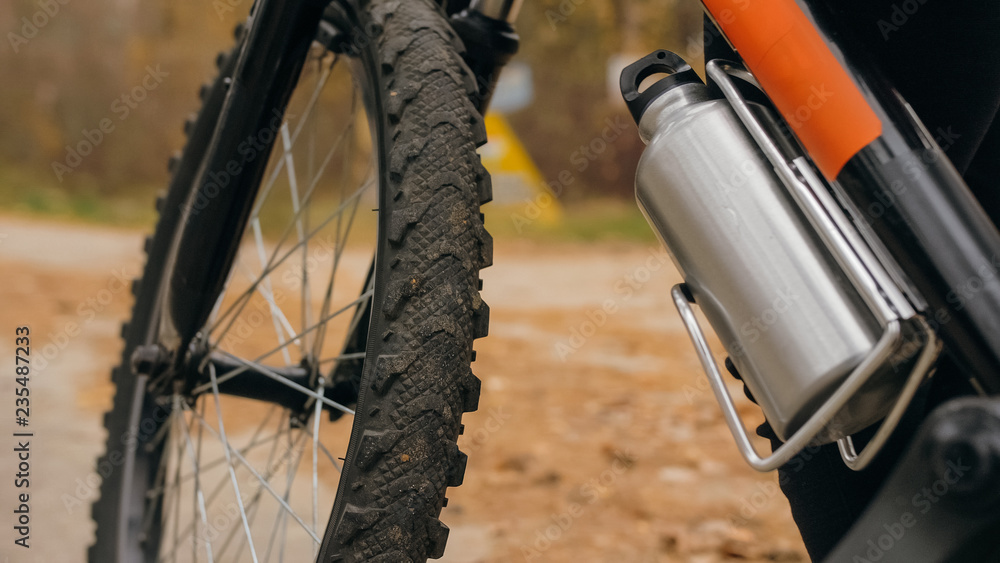 Fototapeta premium Kid drink water from an aluminum flask bottle. One caucasian children rides bike road in autumn park. Little girl riding black orange mtb cycle in forest. Biker motion ride with backpack and helmet.