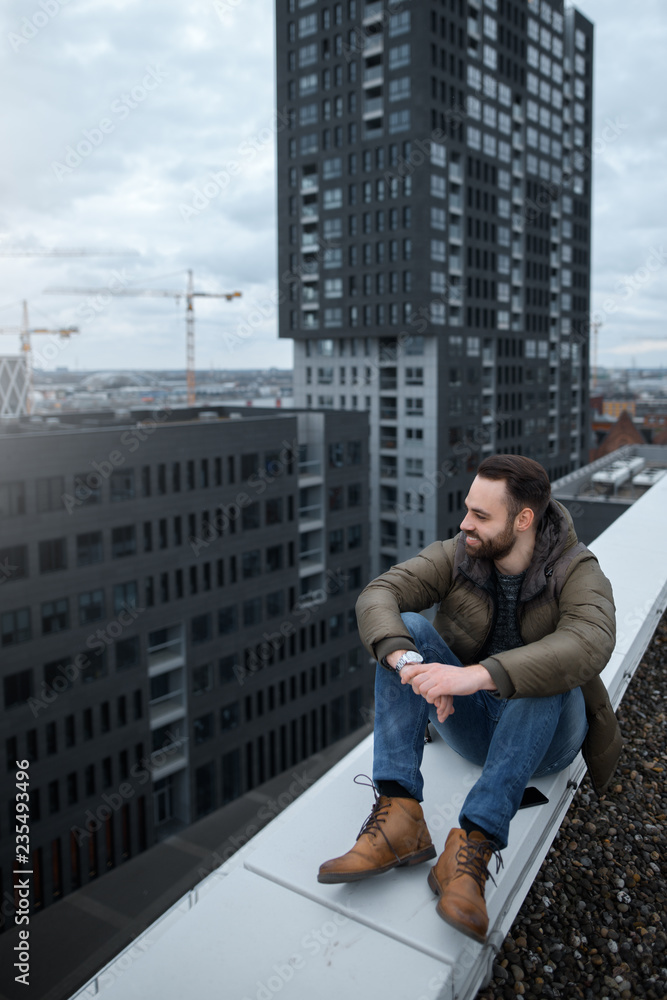 Happy bearded hipster looking at the city from roof of building. Happy bearded hipster looking at the city from roof of building.