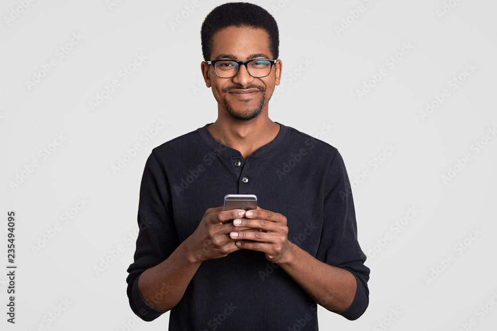 Waist up shot of pleased cheerful dark skinned young man holds smart phone in hands, types message, makes online shopping, connected to wireless internet, being addicted to modern technologies Waist up shot of pleased cheerful dark skinned young man holds smart phone in hands, types message, makes online shopping, connected to wireless internet, being addicted to modern technologies
