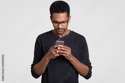 Photo of concentrated dark skinned handsome guy reads news in internet, holds cell phone, wears transparent glasses and sweater, isolated over white background, checks email box, uses free internet © sementsova321