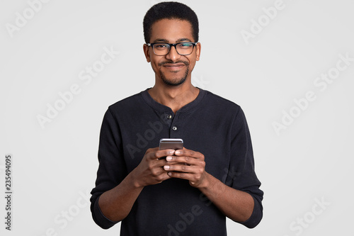 Waist up shot of pleased cheerful dark skinned young man holds smart phone in hands, types message, makes online shopping, connected to wireless internet, being addicted to modern technologies © sementsova321