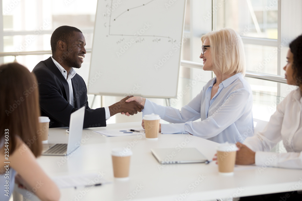 Friendly young african american businessman and smiling mature caucasian businesswoman shaking hands over office table, diverse multiracial partners handshaking at business meeting, respect concept Friendly young african american businessman and smiling mature caucasian businesswoman shaking hands over office table, diverse multiracial partners handshaking at business meeting, respect concept