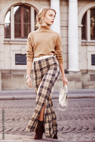 Outdoor full body fashion portrait of young beautiful fashionable girl wearing beige cashmere turtleneck, high-waisted checkered trousers, belt, wrist watch, heels, holding bag, walking in street