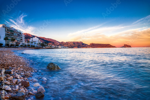Long Exposure of the rocky shore and blue water of Altea Spain with buldings and mountains in the distance and the orange painted sky from the setting sun mixed with blue photo