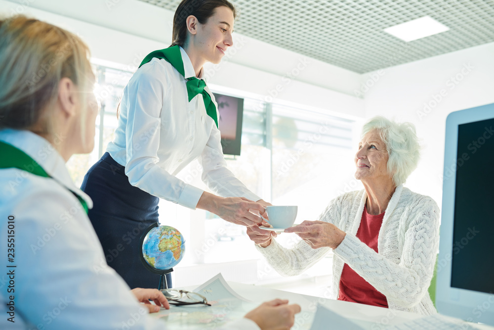Fototapeta premium Content attractive secretary in neck scarf giving cup of coffee to senior lady who choosing travel tour with manager, staff of company taking care of client in travel agency office